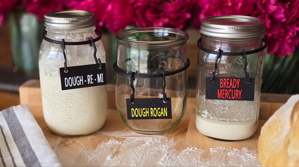 Three glass jars with metallic lids on a wooden surface, containing different types of flour with humorous labels.